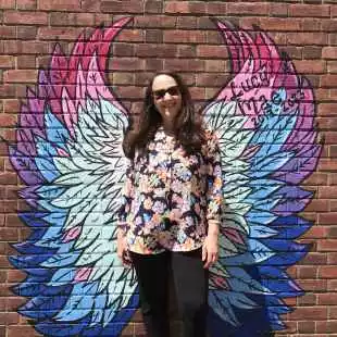 Melissa Brown in front of a brick wall with painted bright colored angel wings behind her