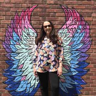 Melissa Brown in front of a brick wall with painted bright colored angel wings behind her