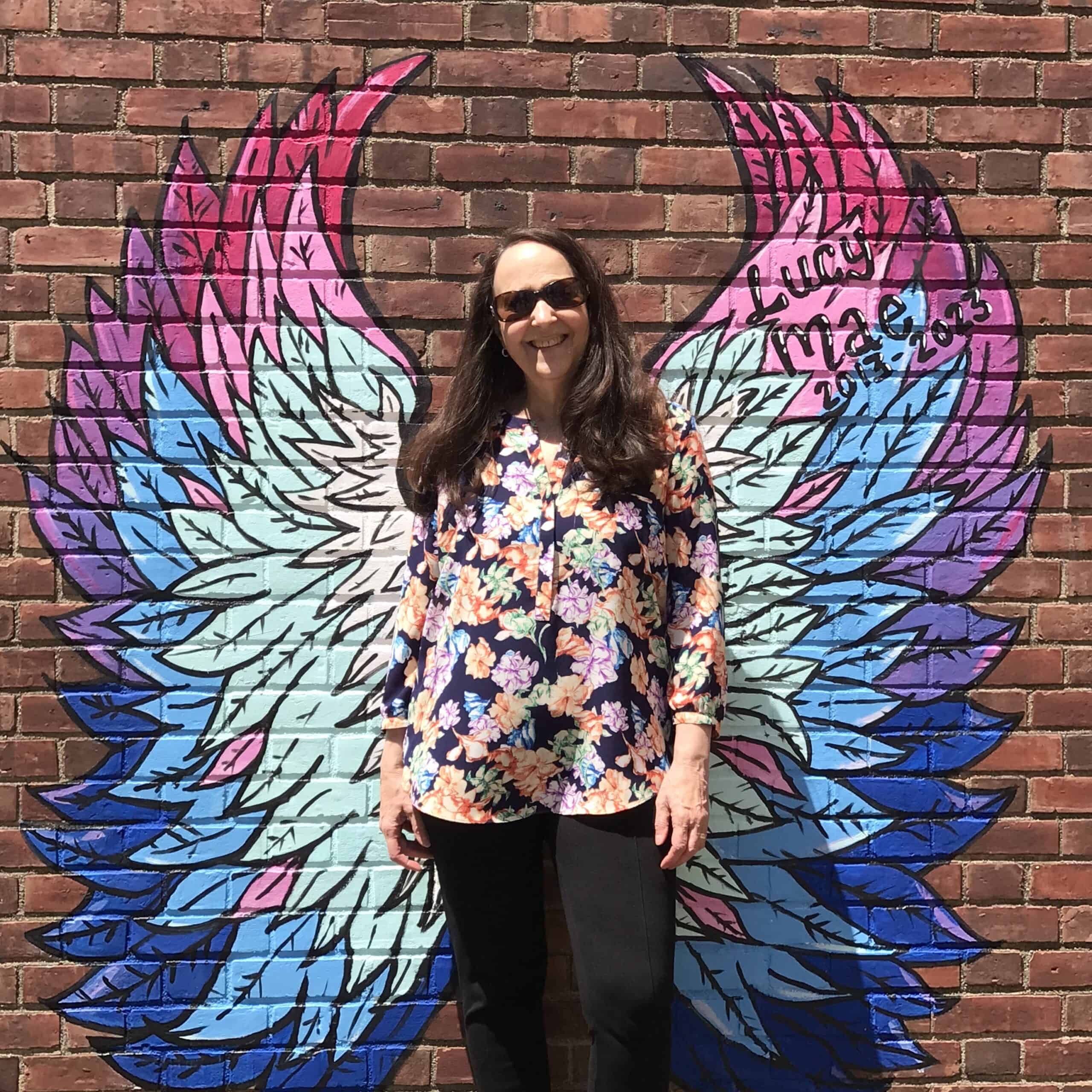 Melissa Brown in front of a brick wall with painted bright colored angel wings behind her