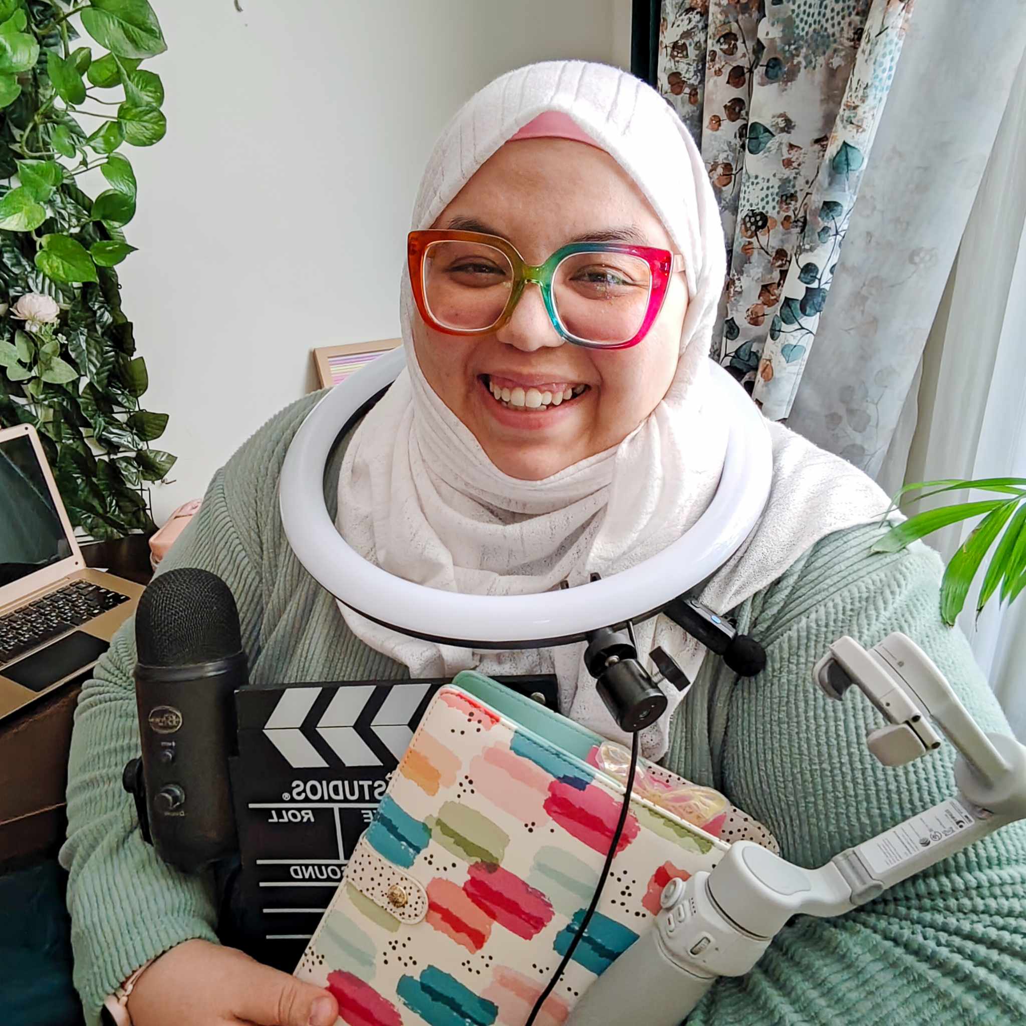 Woman with headscarf smiling while holding a notebook and podcasting equipment.