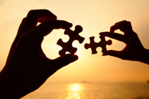 Two hands holding jigsaw puzzle pieces aligning to connect against a sunset background.