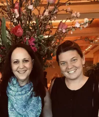 Two women smiling at the camera, one wearing a blue scarf, standing in front of a floral arrangement indoors.