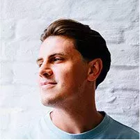 A smiling woman with curly red-tinted hair, wearing a blue blazer and a purple top paired with a pearl necklace, posing in front of a brick building.